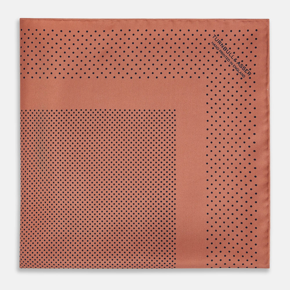 The Turnbull & Asser Blush Dot Silk Pocket Square features a rust hue with black polka dots, a bordered design, and hand-rolled edges, shown folded against a white background.
