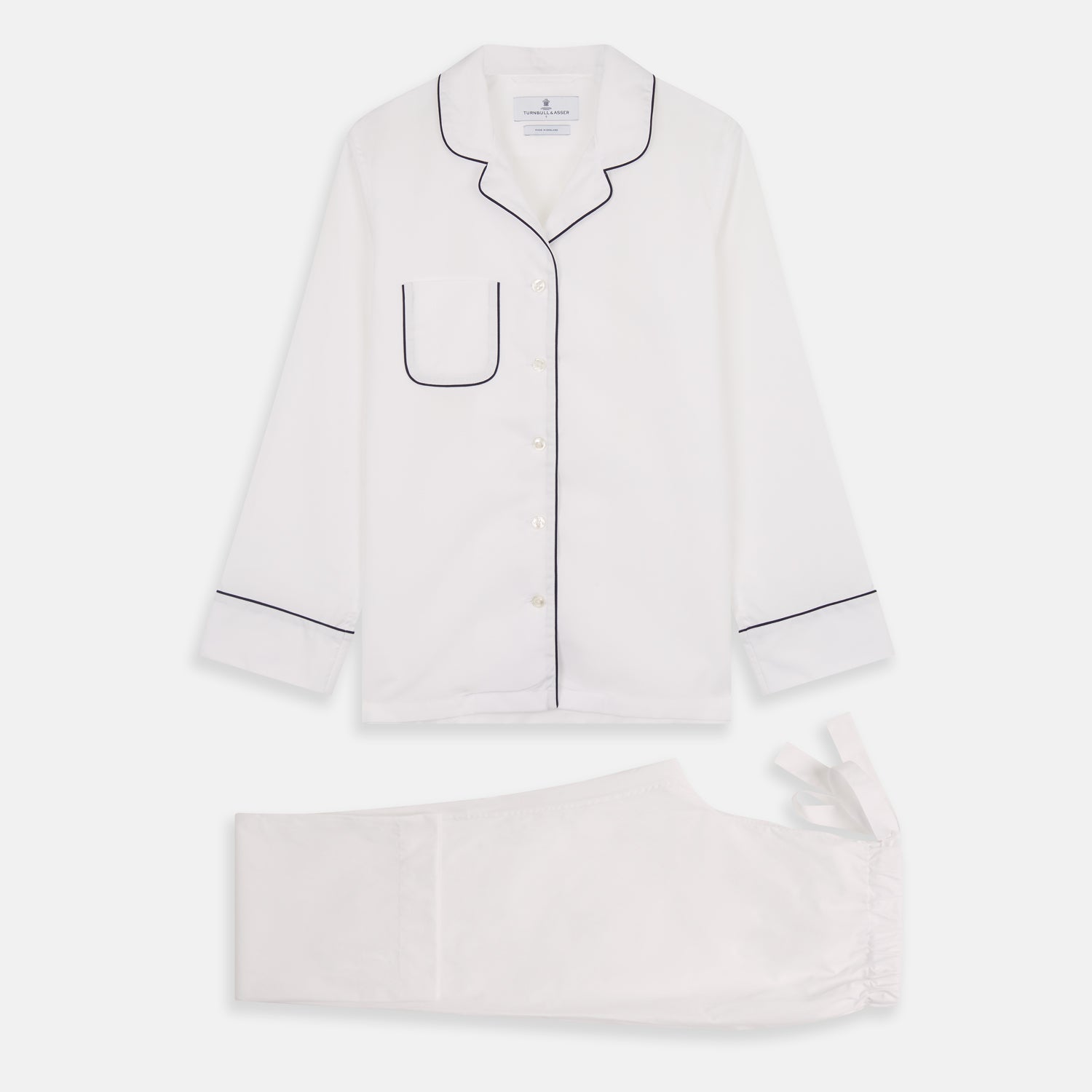 View 6 of product The Turnbull & Asser Women's Harriet Plain White Cotton Pyjama Set features long sleeves, black piping, a chest pocket, and matching pants with elastic waistband and ribbon tie, all displayed on a white background.