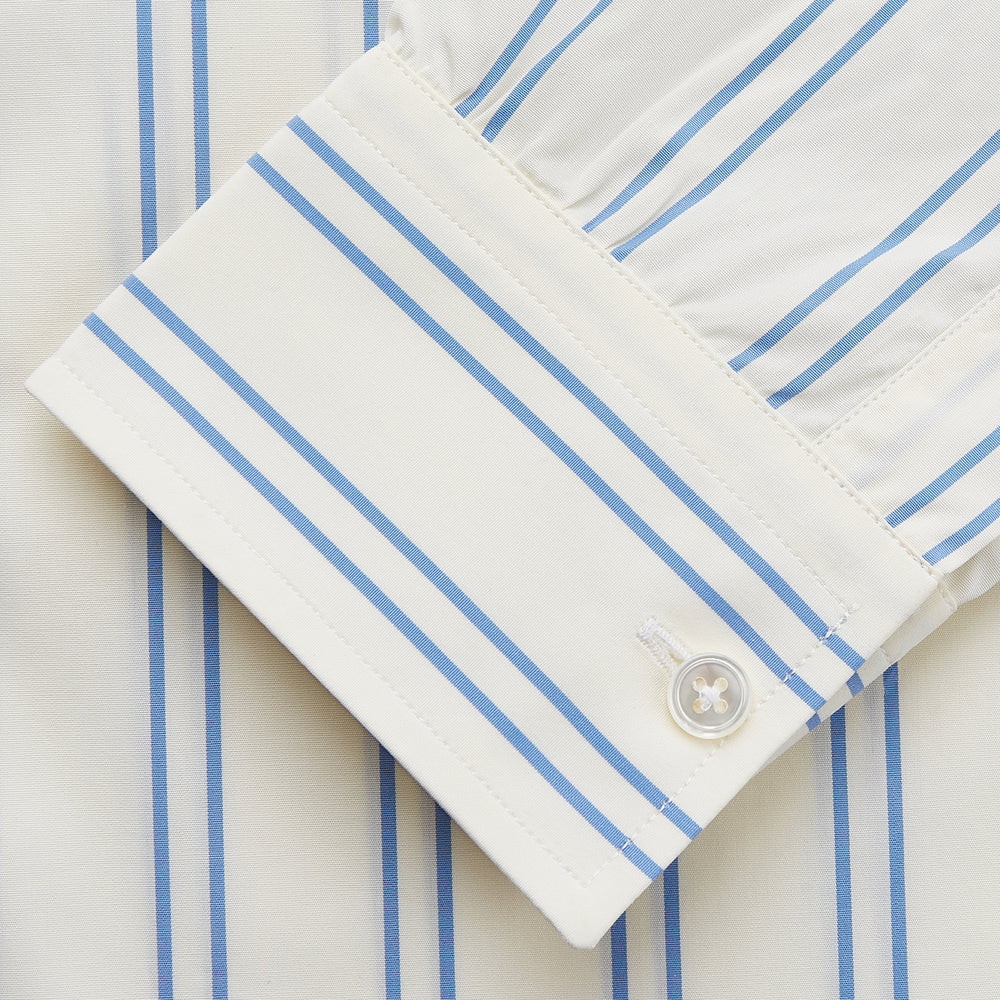 A man with short brown hair stands against a beige background in a Turnbull & Asser Blue and Off-White Stripe Cotton Weekend Fit Finch Shirt, featuring vertical stripes and a Derby collar, paired with dark pants. One hand is in his pocket; he has a neutral expression.