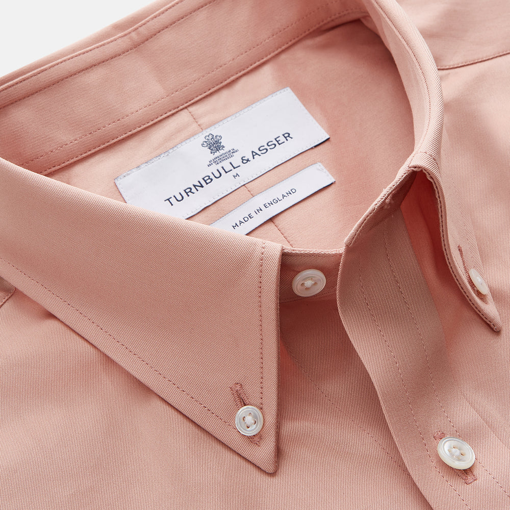 A man faces forward in a relaxed pose against a beige background, wearing the Turnbull & Asser Pink Piece Dyed Weekend Fit Suffolk Shirt with a button-down collar, dark cuffed jeans, and black shoes.