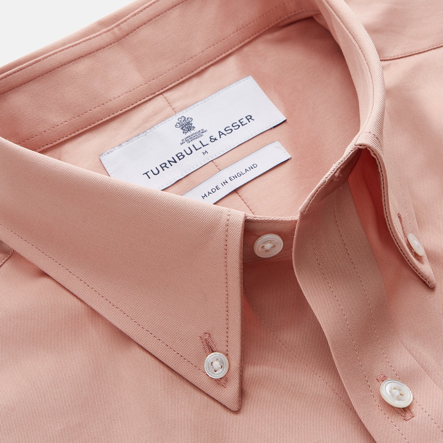View 3 of product A man faces forward in a relaxed pose against a beige background, wearing the Turnbull & Asser Pink Piece Dyed Weekend Fit Suffolk Shirt with a button-down collar, dark cuffed jeans, and black shoes.