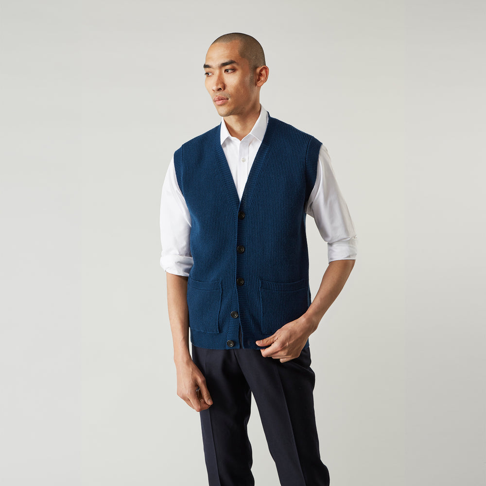 A man with a shaved head wears a Turnbull & Asser Navy Sleeveless Cashmere Cardigan over a white dress shirt with rolled-up sleeves and dark trousers, standing against a plain light gray background.