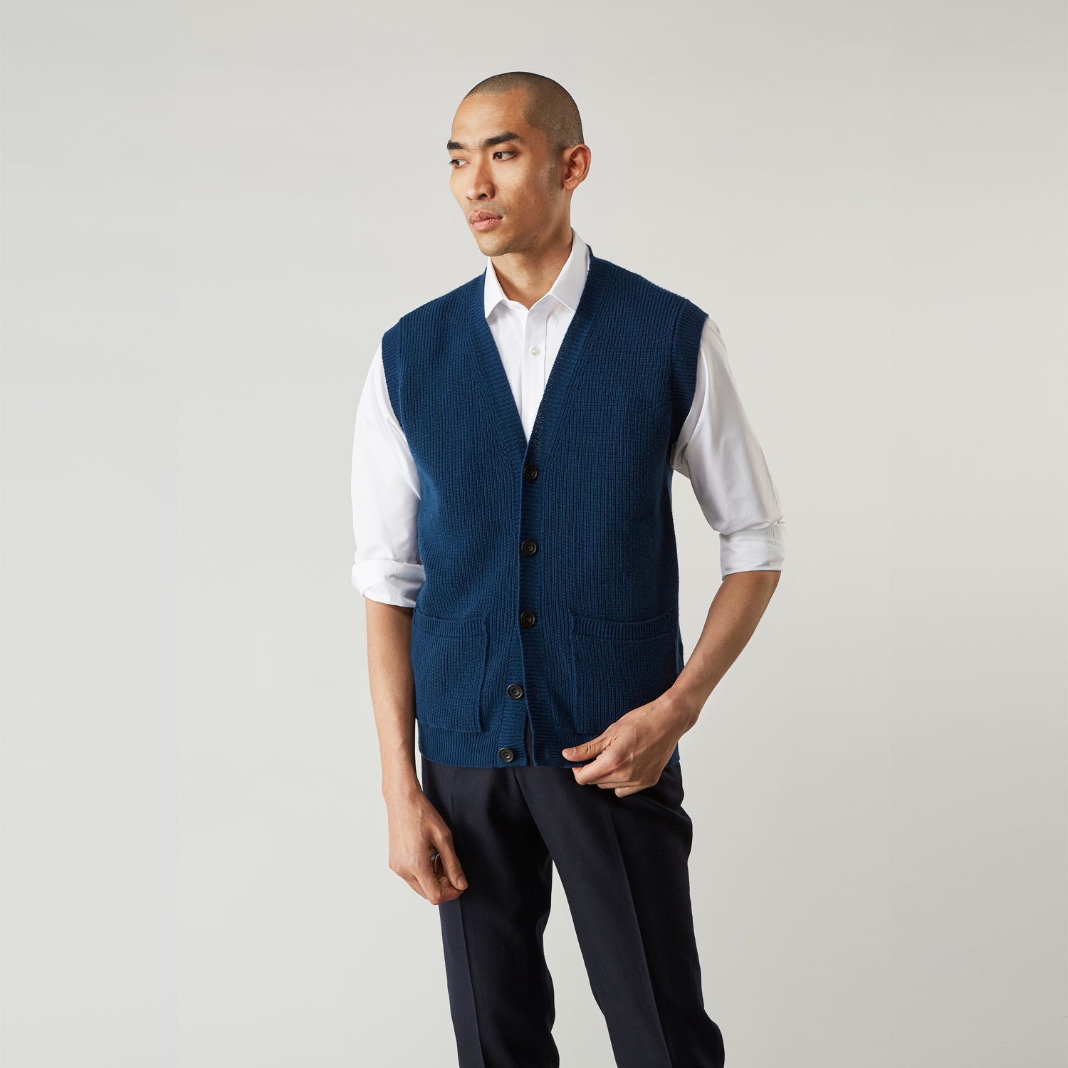 View 1 of product A man with a shaved head wears a Turnbull & Asser Navy Sleeveless Cashmere Cardigan over a white dress shirt with rolled-up sleeves and dark trousers, standing against a plain light gray background.