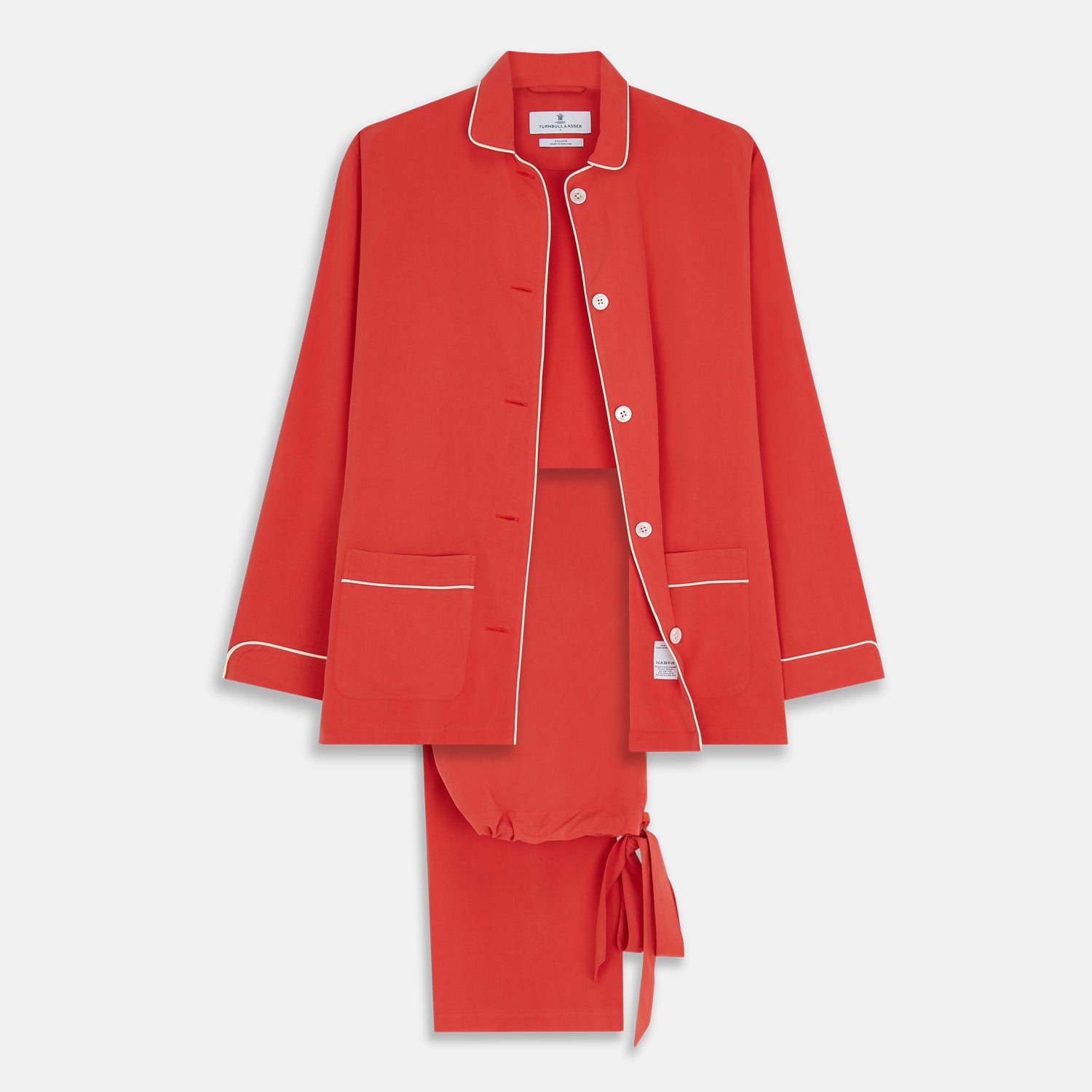 View 1 of product The Turnbull & Asser Women's Coral Pink Silk Thelma Pyjama Set features a long-sleeve button-up top with two front pockets and matching drawstring pants, shown on a white background.