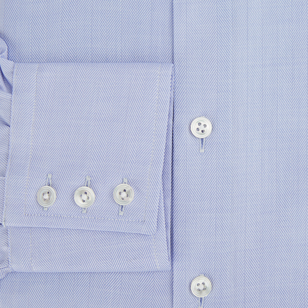 The Turnbull & Asser BLUE HERRINGBONE TAILORED FIT SHIRT WITH KENT COLLAR features long sleeves and buttoned cuffs, offering a timeless formal look in a classic light blue herringbone fabric, laid flat on a white background.