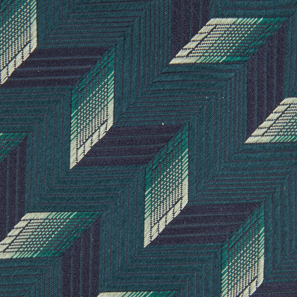 The Turnbull & Asser "The Dark Knight Silk Tie as seen on the Joker" is shown rolled up, featuring a teal zigzag geometric pattern with blue, green, and cream diamonds on a white background.