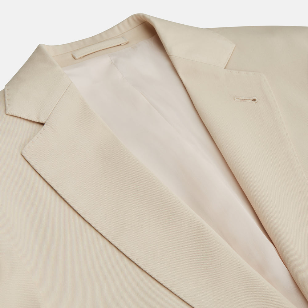 A man in a Turnbull & Asser Beige Cotton Barrington Blazer, light yellow shirt, blue patterned tie, and dark trousers stands against a plain light background, with a pocket square visible and a neutral expression.