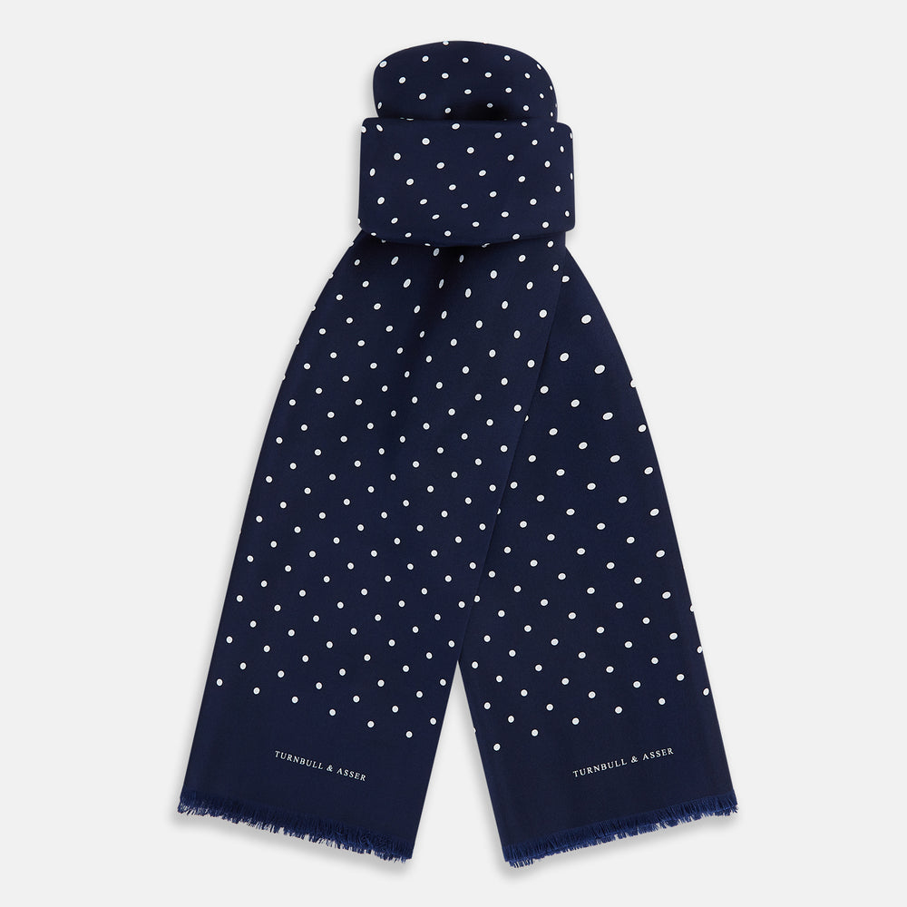 The Navy and White Spotted Silk Scarf by Turnbull & Asser features white polka dots, a fringed edge, and the brand's name printed near the bottom on both sides. It is neatly folded at the top.