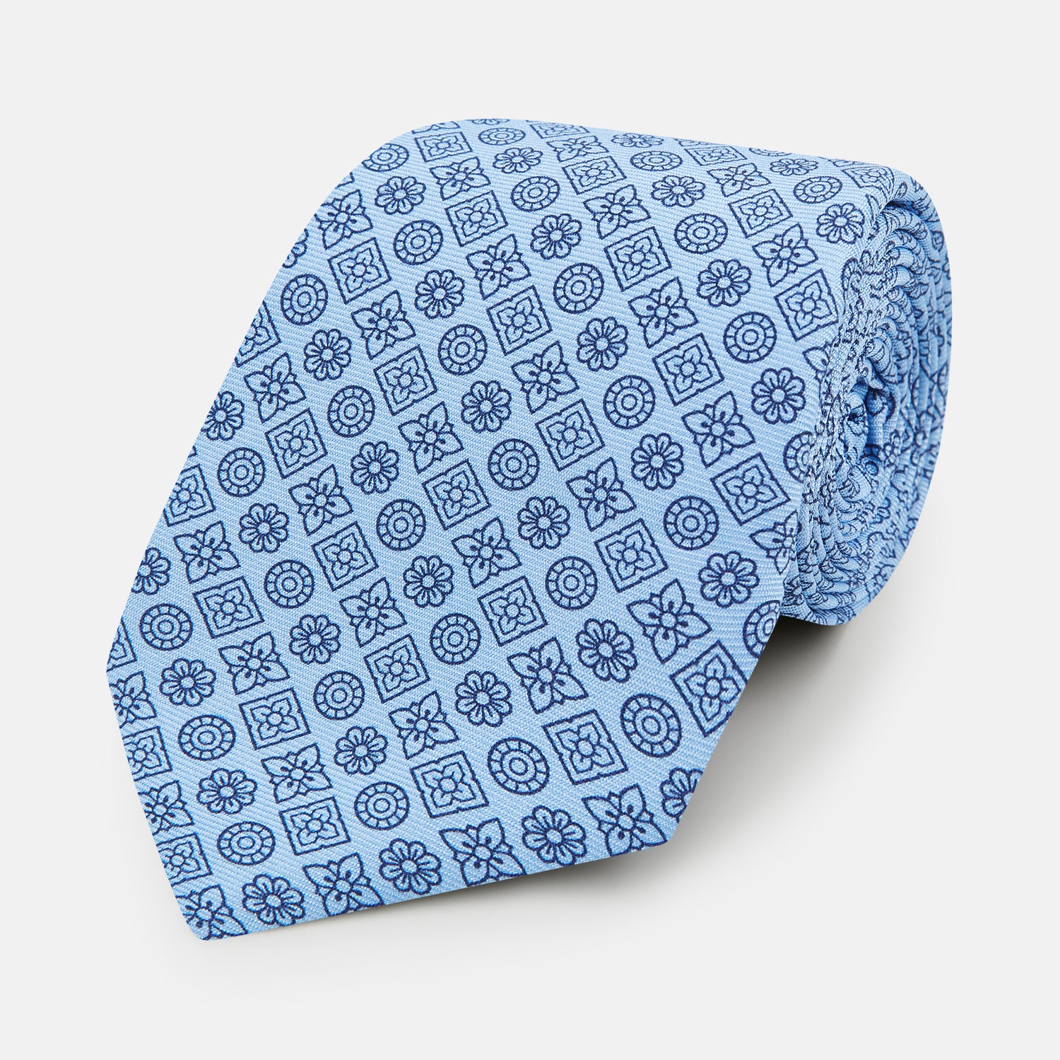 View 1 of product The Turnbull & Asser Blue Floral Tile Pattern Printed Silk Tie features a light blue geometric and floral design in dark blue, shown rolled up on a white background.