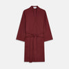 View 2 of product A person stands indoors by a window, wearing the Turnbull & Asser Burgundy Cotton-Cashmere Kimono Gown over striped pajamas, with matching slippers, next to cream curtains and a wooden chair against a pale wall.