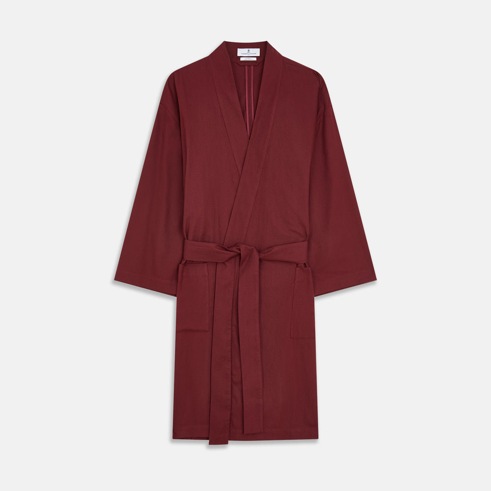 A person stands indoors by a window, wearing the Turnbull & Asser Burgundy Cotton-Cashmere Kimono Gown over striped pajamas, with matching slippers, next to cream curtains and a wooden chair against a pale wall.