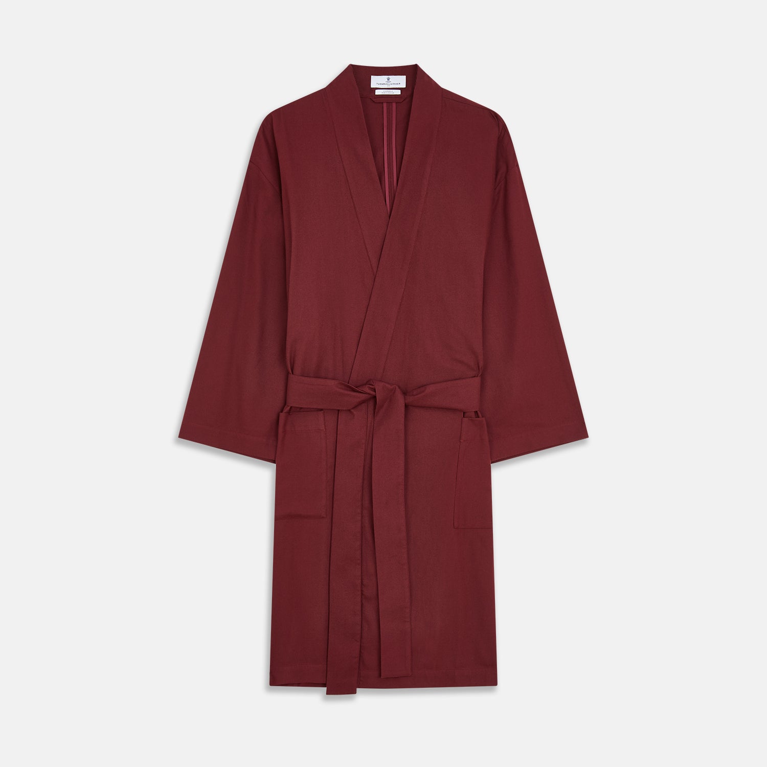 View 2 of product A person stands indoors by a window, wearing the Turnbull & Asser Burgundy Cotton-Cashmere Kimono Gown over striped pajamas, with matching slippers, next to cream curtains and a wooden chair against a pale wall.
