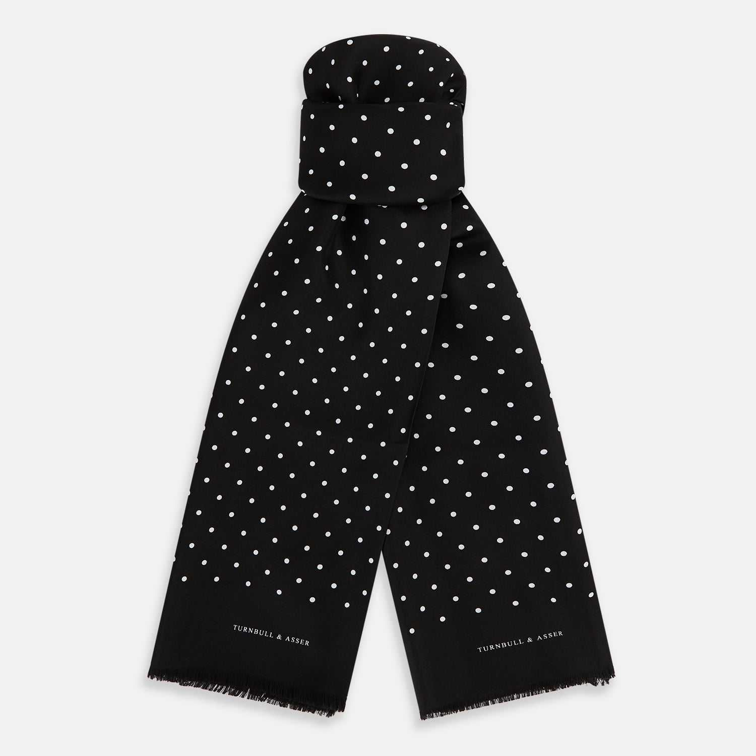 View 1 of product The Black and White Spotted Silk Scarf by Turnbull & Asser features a spotted design, neatly folded at the top and spread out at the bottom, with the brand's name printed in white near its fringed ends.