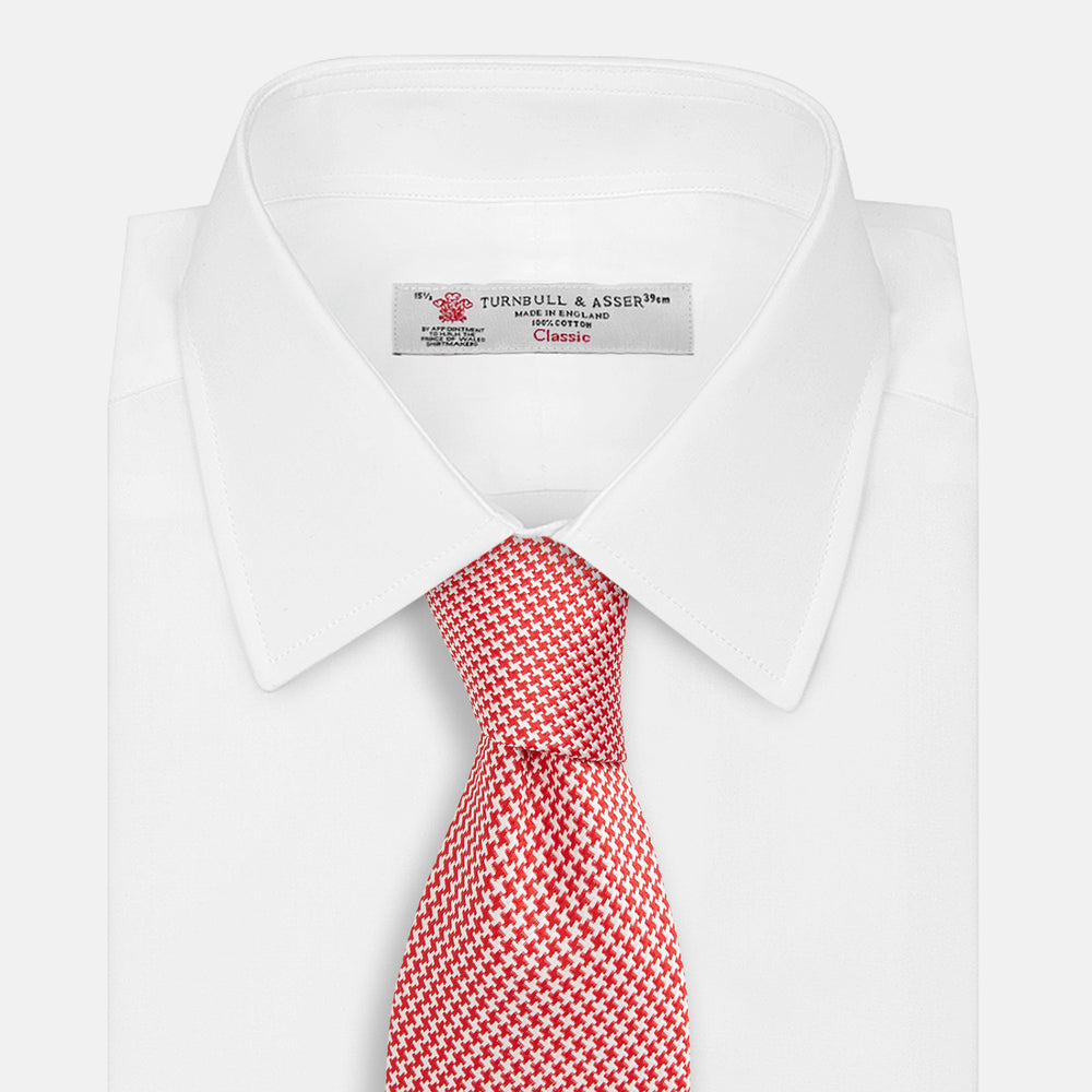 The Turnbull & Asser Red and White patterned silk tie, crafted from fine silk with a small geometric design, is elegantly rolled and displayed on a white background—a refined men’s accessory.