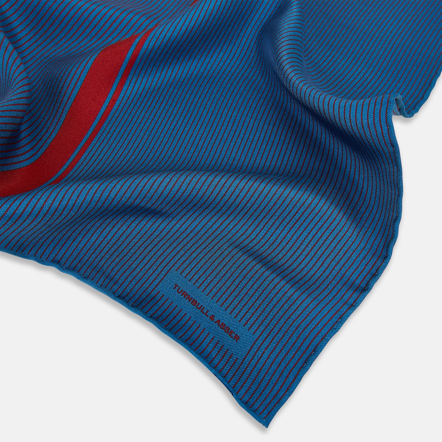 View 2 of product A blue silk pocket square by Turnbull & Asser, featuring thin black stripes, a striking vertical red stripe, and a turned-over corner that reveals the reverse side. The brand name is printed discreetly in one corner.