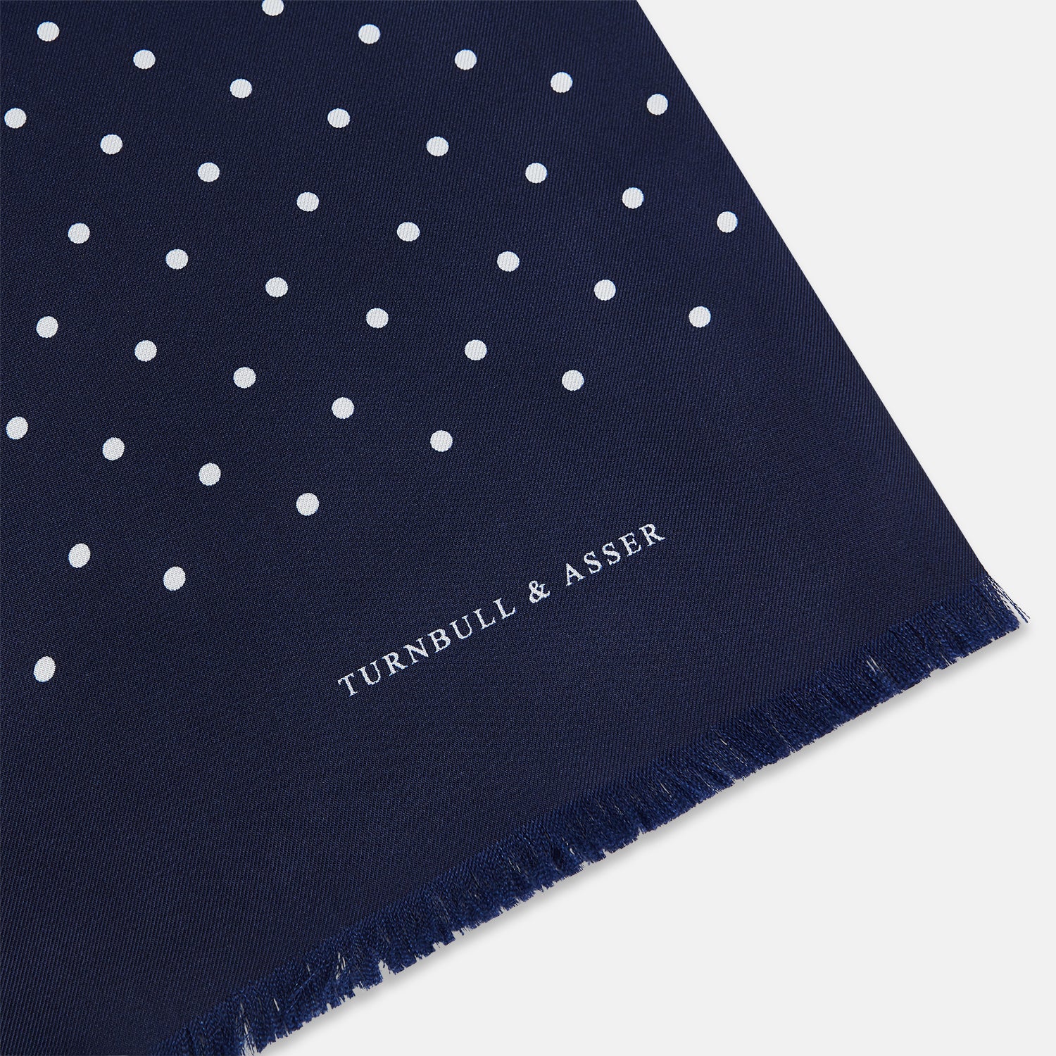View 2 of product The Navy and White Spotted Silk Scarf by Turnbull & Asser features white polka dots, a fringed edge, and the brand's name printed near the bottom on both sides. It is neatly folded at the top.