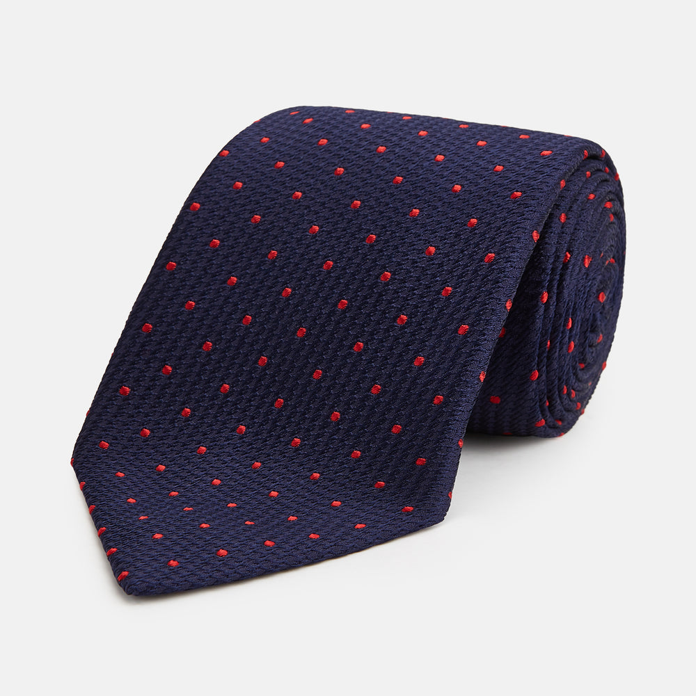 The Turnbull & Asser LACE ON TONE SPOT NAVY TULIP RED TIE is a classic men's accessory crafted from textured navy fabric with small red polka dots, neatly rolled with the pointed end facing forward.