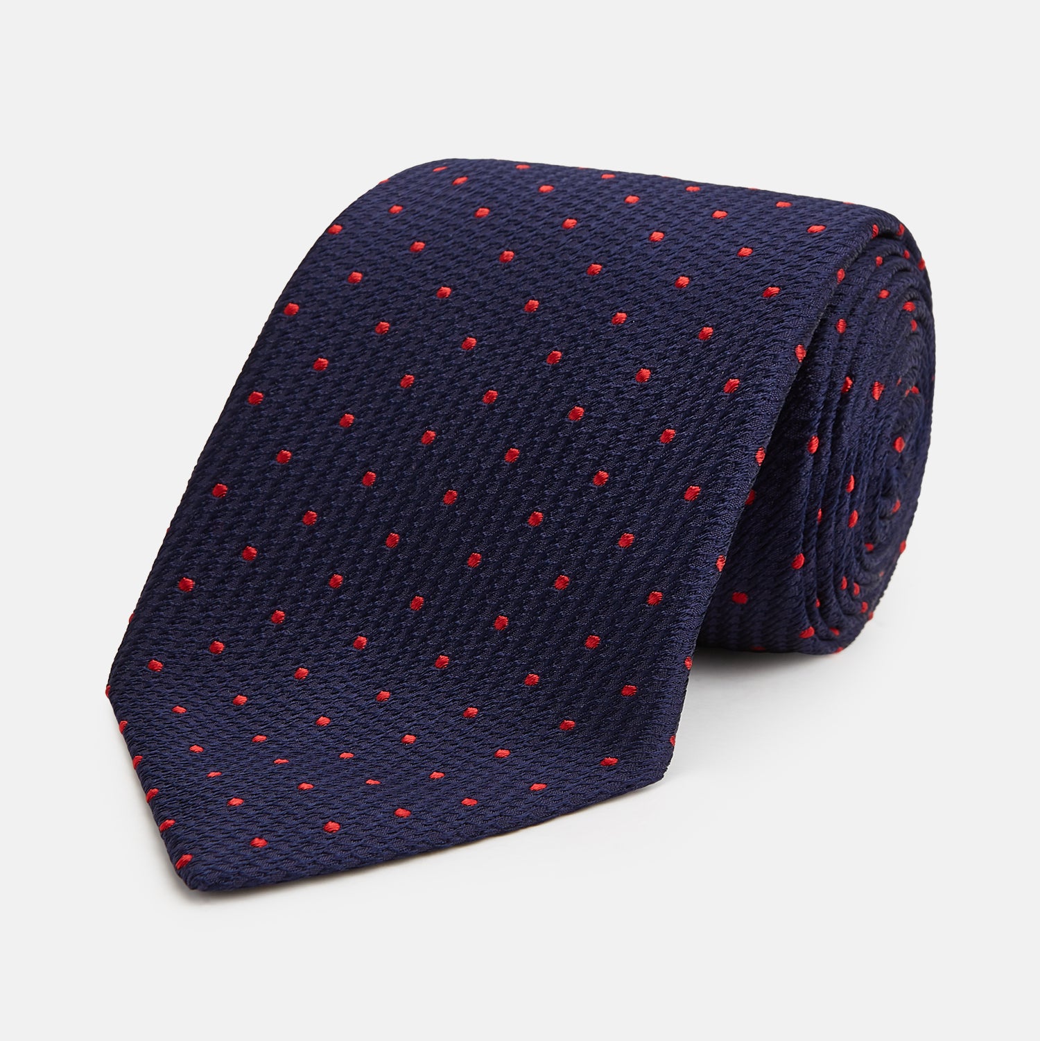 View 1 of product The Turnbull & Asser LACE ON TONE SPOT NAVY TULIP RED TIE is a classic men's accessory crafted from textured navy fabric with small red polka dots, neatly rolled with the pointed end facing forward.