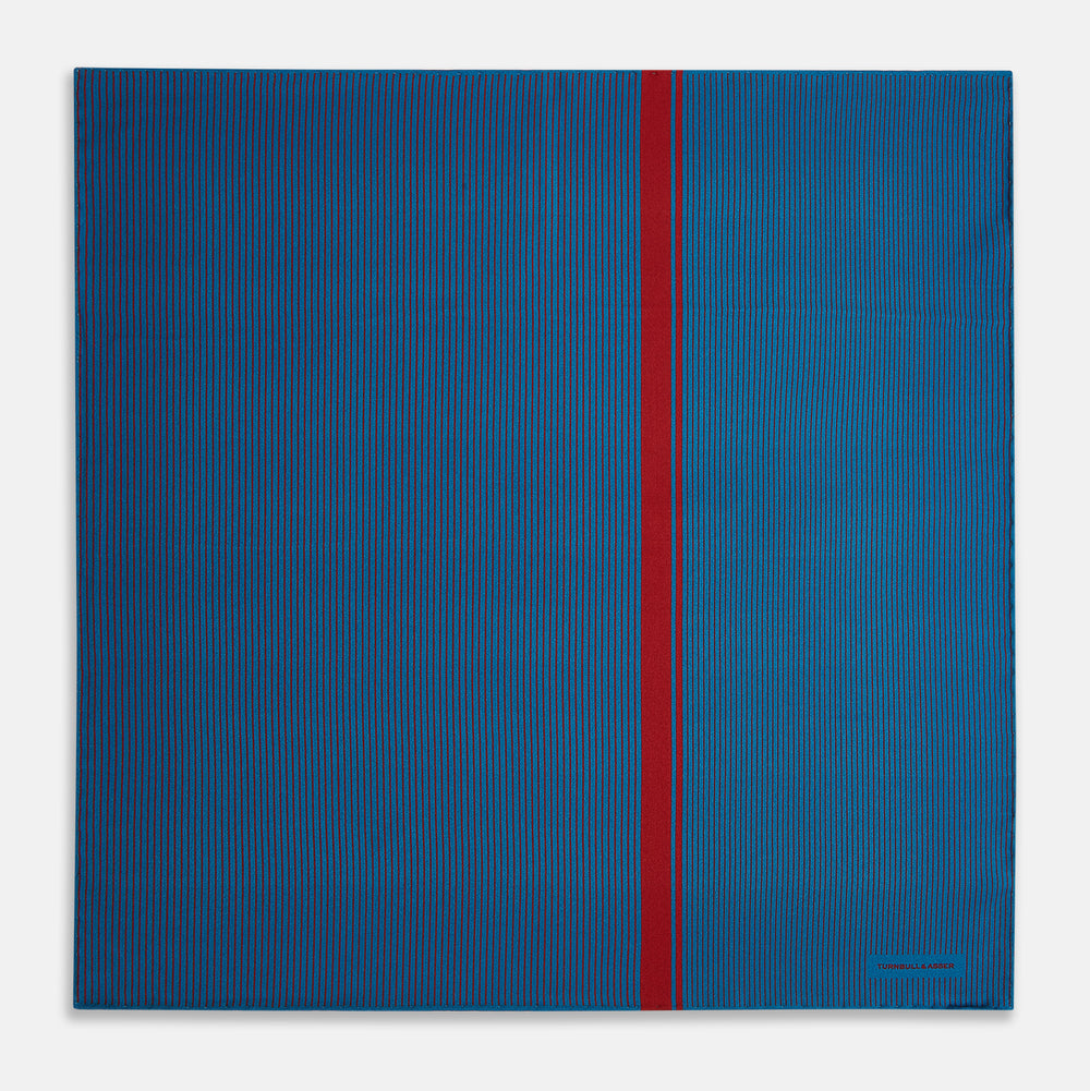 A blue silk pocket square by Turnbull & Asser, featuring thin black stripes, a striking vertical red stripe, and a turned-over corner that reveals the reverse side. The brand name is printed discreetly in one corner.