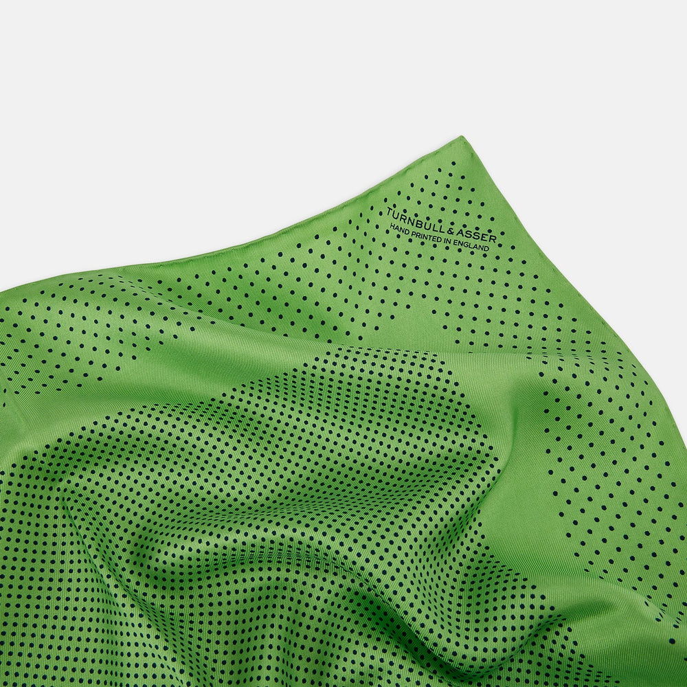 The Fern Green Dot Silk Pocket Square by Turnbull & Asser is crafted from pure silk, featuring a subtle black polka dot border with a denser dot pattern at the center and the brand name printed in the upper right corner.
