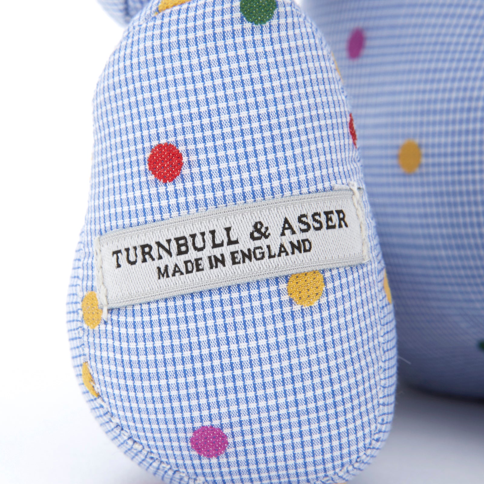 View 3 of product The Turnbuline Cotton Spot Teddy Bear by Turnbull & Asser is a blue gingham bear with colorful polka dots, black button eyes, a black nose, and a white leg label, shown sitting upright in profile facing left.