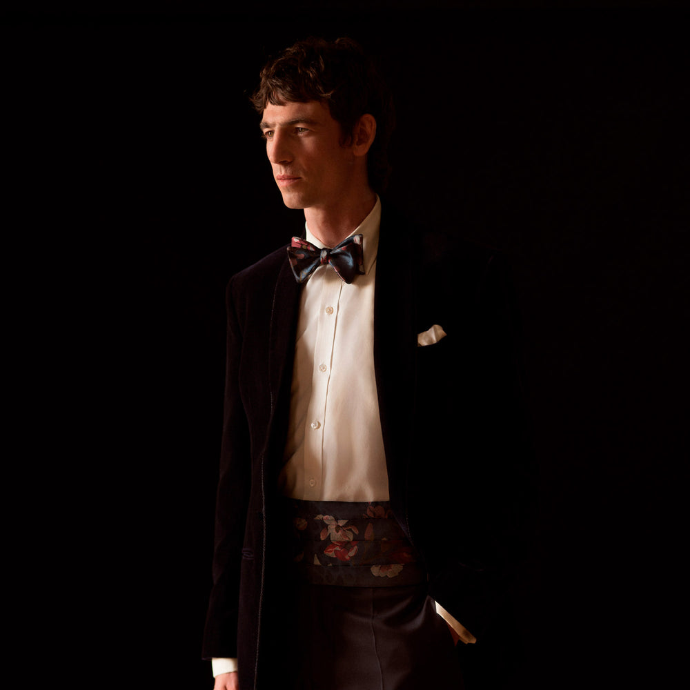 A man wearing a Turnbull & Asser Exclusive Silk Magnolia Cummerbund, paired with a black tuxedo jacket, white dress shirt, and patterned bow tie, stands against a dark background with a serious look.