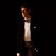 A man wearing a Turnbull & Asser Exclusive Silk Magnolia Cummerbund, paired with a black tuxedo jacket, white dress shirt, and patterned bow tie, stands against a dark background with a serious look.