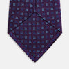 View 4 of product The Turnbull & Asser MINI SQUARE SPOT PLUM BLUE TIE is a luxury English silk necktie in plum, featuring a blue and white grid pattern of small squares, displayed rolled up against a simple light background.