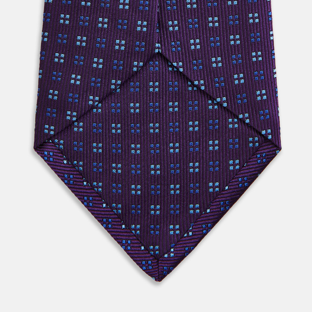 The Turnbull & Asser MINI SQUARE SPOT PLUM BLUE TIE is a luxury English silk necktie in plum, featuring a blue and white grid pattern of small squares, displayed rolled up against a simple light background.