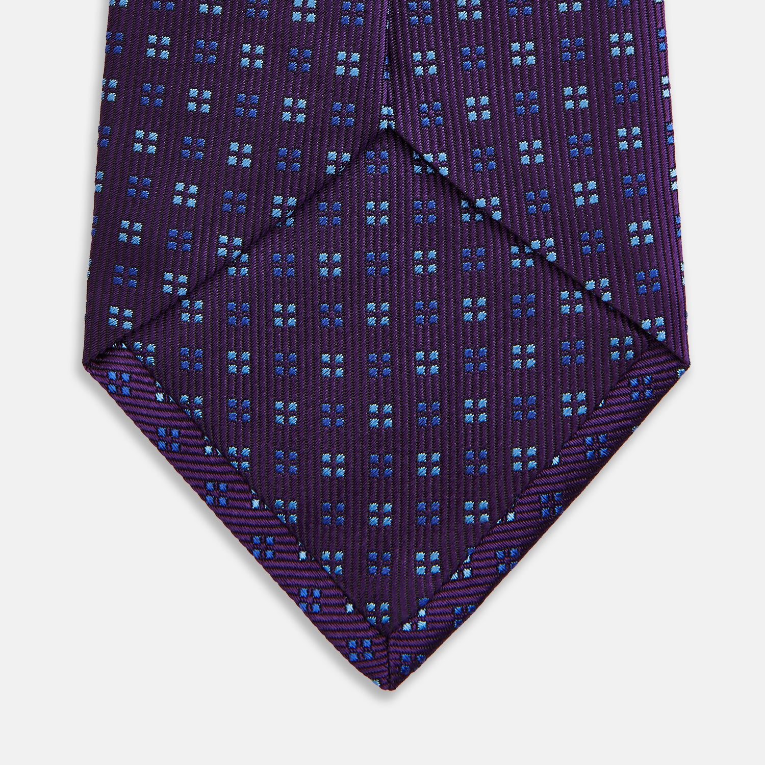 View 4 of product The Turnbull & Asser MINI SQUARE SPOT PLUM BLUE TIE is a luxury English silk necktie in plum, featuring a blue and white grid pattern of small squares, displayed rolled up against a simple light background.