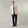 View 3 of product A man in a Turnbull & Asser Beige Cotton Barrington Blazer, light yellow shirt, blue patterned tie, and dark trousers stands against a plain light background, with a pocket square visible and a neutral expression.