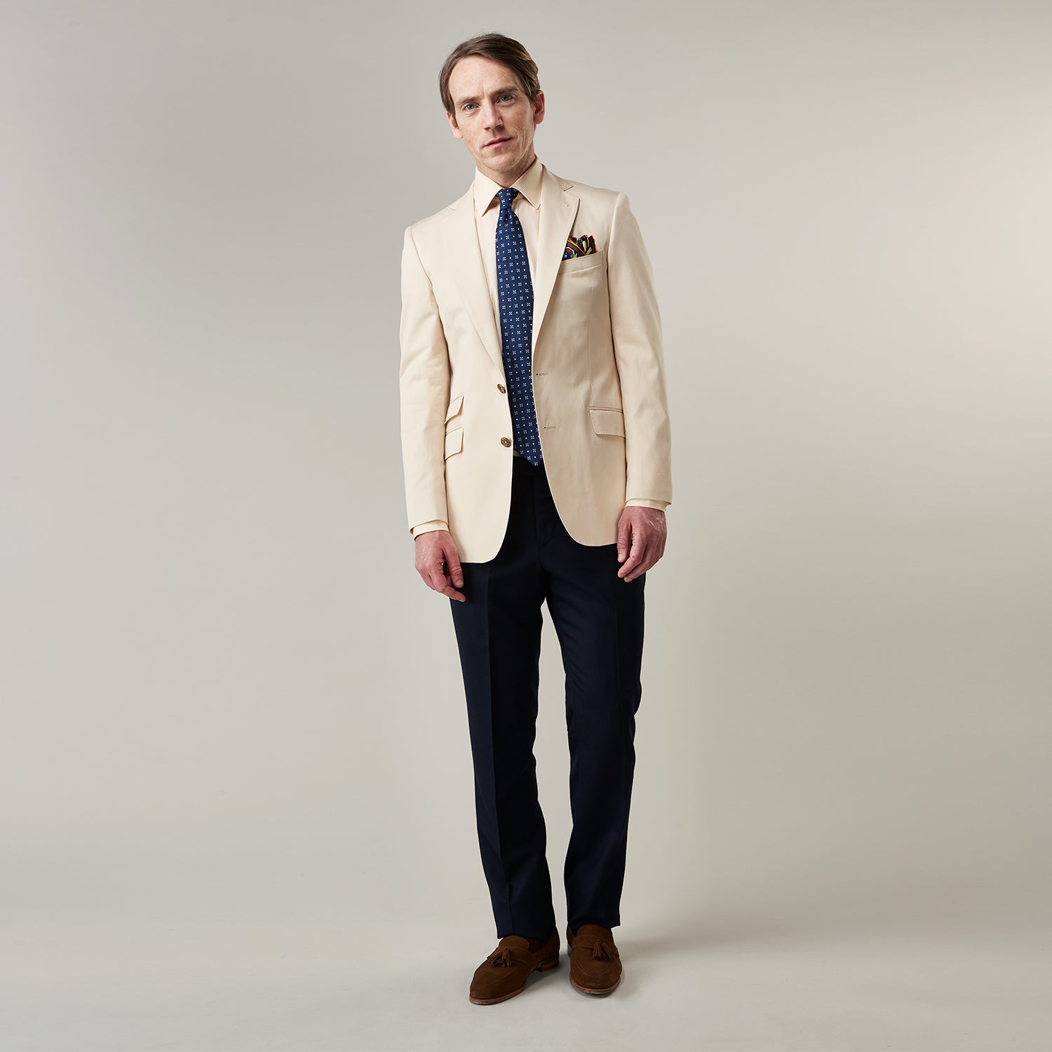 View 3 of product A man in a Turnbull & Asser Beige Cotton Barrington Blazer, light yellow shirt, blue patterned tie, and dark trousers stands against a plain light background, with a pocket square visible and a neutral expression.