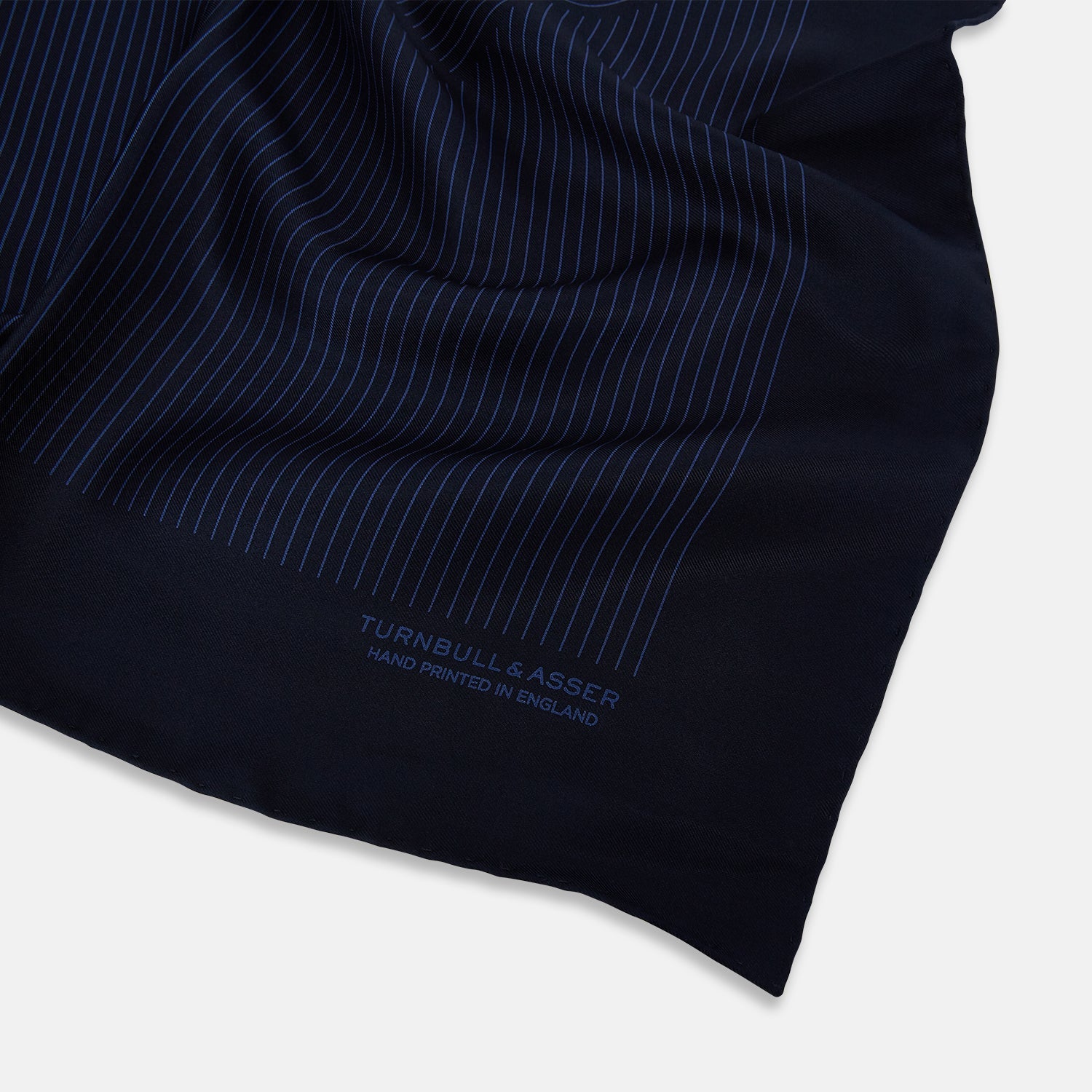 View 2 of product The Navy Stripe Silk Pocket Square by Turnbull & Asser features a dark navy silk with thin light blue stripes and hand-rolled edges, folded to reveal both sides and “TURNBULL & ASSER HAND PRINTED IN ENGLAND” in the corner.