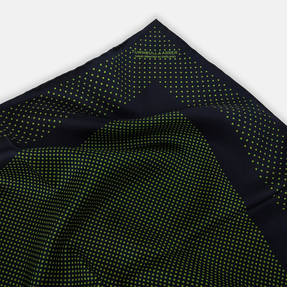 The Turnbull & Asser Navy and Kelly Green Dot Silk Pocket Square features small green dots on navy silk and a folded corner displaying the reverse—an elegant, handmade accessory perfect for enhancing any formal attire.