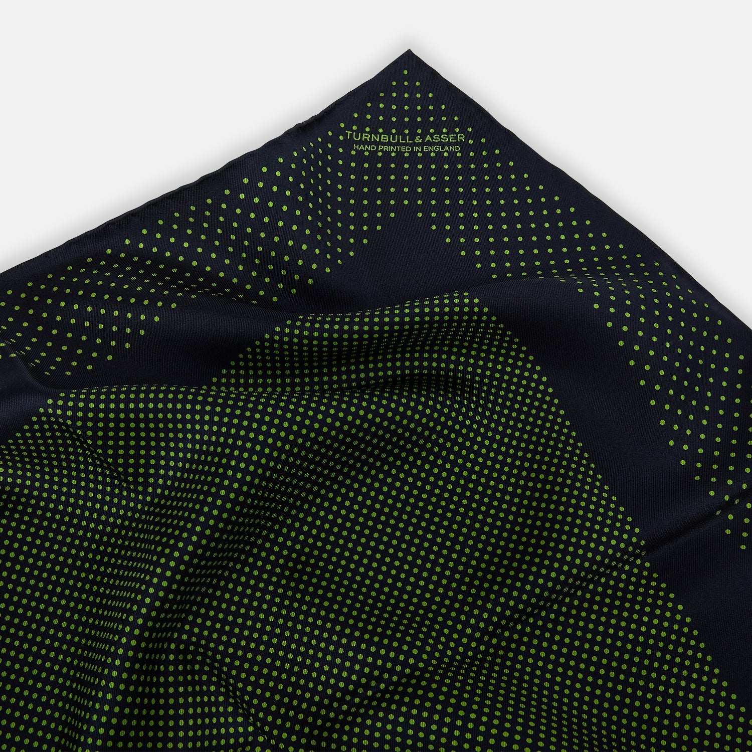View 2 of product The Turnbull & Asser Navy and Kelly Green Dot Silk Pocket Square features small green dots on navy silk and a folded corner displaying the reverse—an elegant, handmade accessory perfect for enhancing any formal attire.