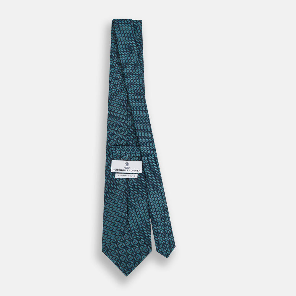 The Turnbull & Asser Turquoise Micro Hexagon Tie features refined British elegance with its small blue hexagonal pattern on a lighter background, neatly rolled and displayed on a plain white surface.