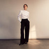 View 5 of product A woman stands in profile, looking down with hands in her pockets, wearing the Turnbull & Asser Turnbull and The Deck White Cotton Expressionist Shirt, paired with high-waisted black-and-white houndstooth pants.