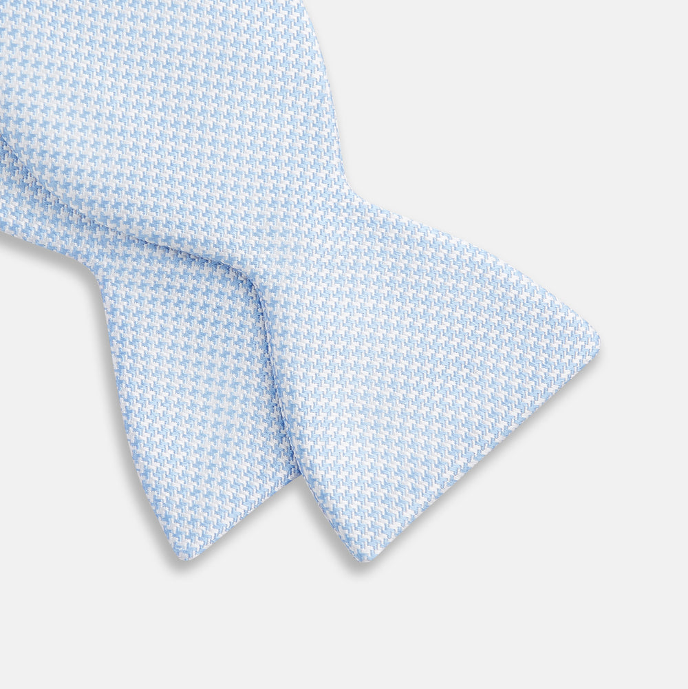 The Turnbull & Asser Pale Blue and White Houndstooth Silk Bow Tie features a refined textured pattern, making it an elegant formalwear accessory against a crisp white background.