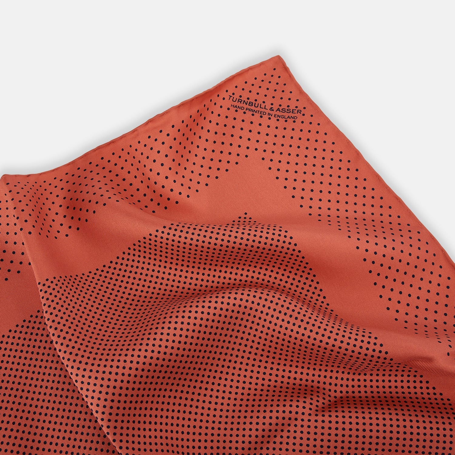 View 2 of product The Orange Dot Silk Pocket Square by Turnbull & Asser is handmade in England from silk, featuring small black polka dots and a solid orange border with a folded corner.