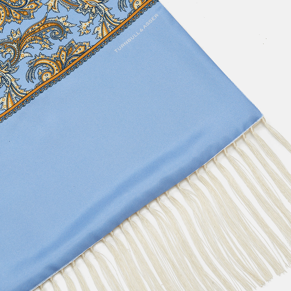 The Turnbull & Asser GGSC3128 5665 06 OS is a blue paisley silk scarf with ornate gold designs, solid light blue ends, and cream fringes. Shown laid flat with one end folded, it highlights its luxurious style.