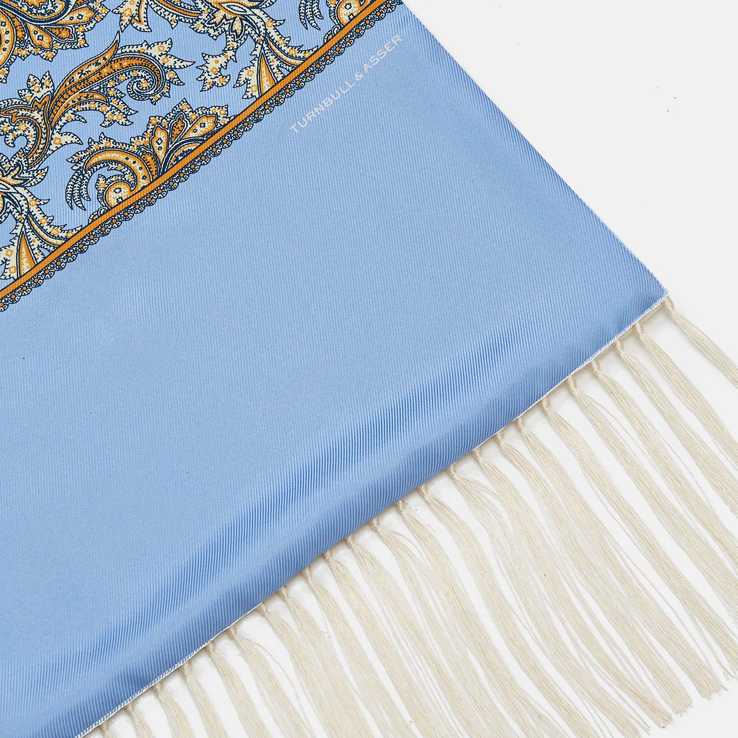 View 2 of product The Turnbull & Asser GGSC3128 5665 06 OS is a blue paisley silk scarf with ornate gold designs, solid light blue ends, and cream fringes. Shown laid flat with one end folded, it highlights its luxurious style.