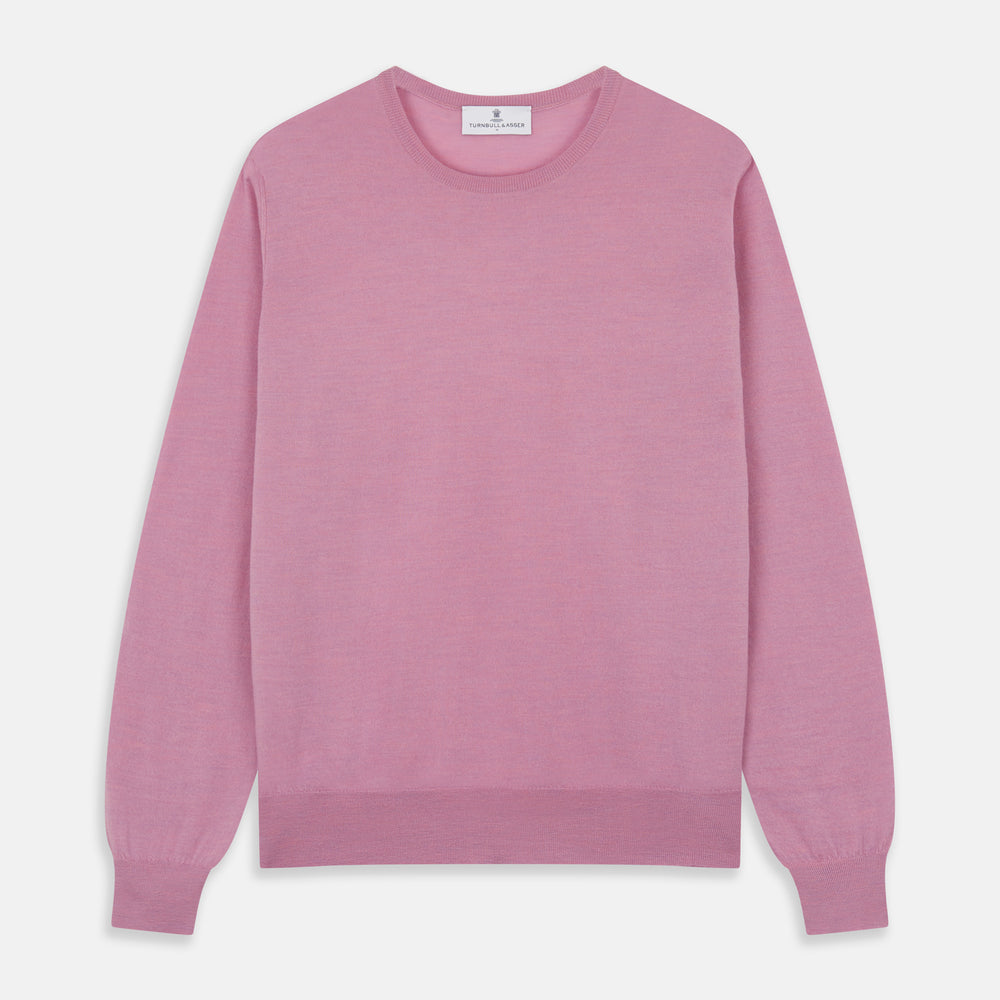 Wearing Turnbull & Asser’s Pink Merino Kingston Crew Neck Jumper, white pants, and a colorful scarf, a person stands against a plain light gray background with hands in their pockets.