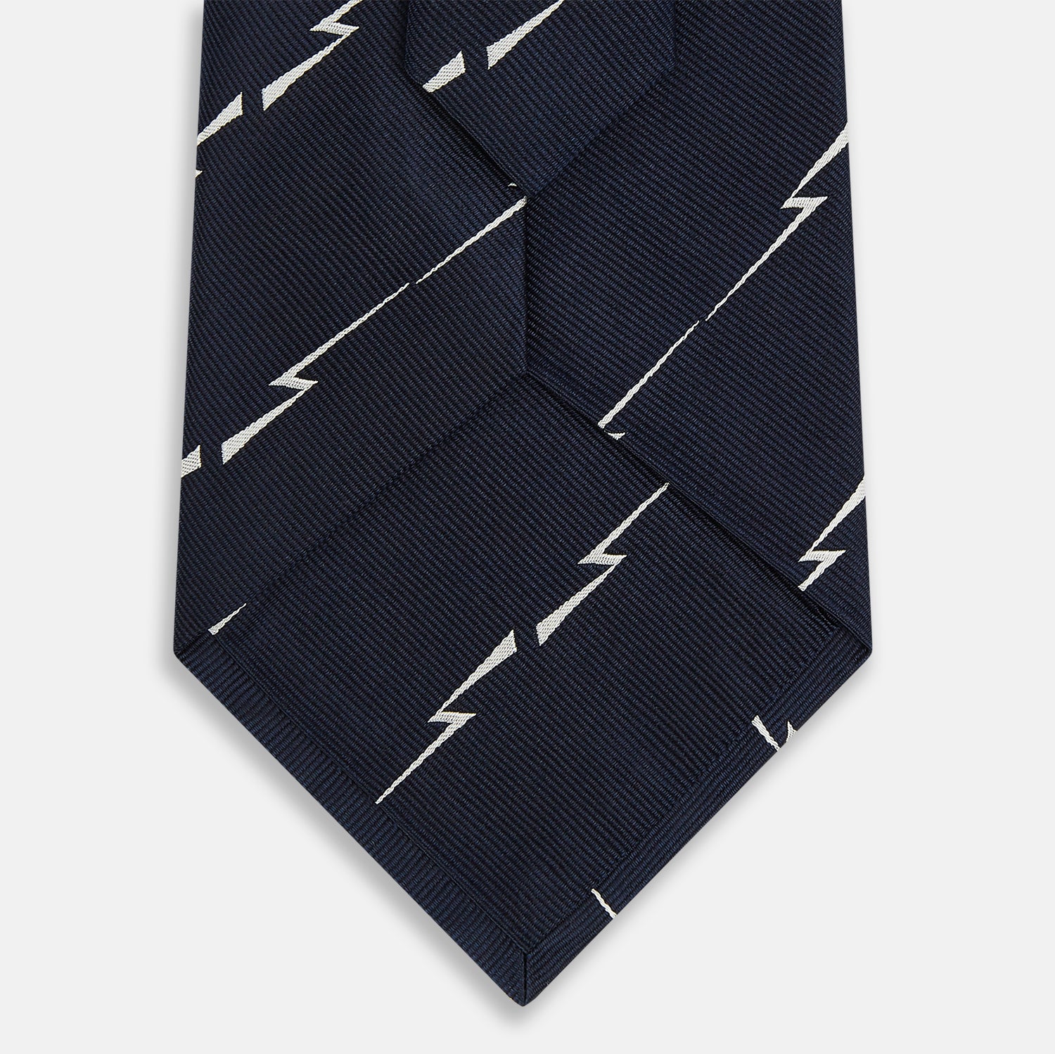 View 3 of product The Turnbull & Asser Navy Zigzag Jacquard Silk Tie is crafted from fine English silk, featuring a navy base with thin white zigzag stripes and shown rolled up on a plain white background.