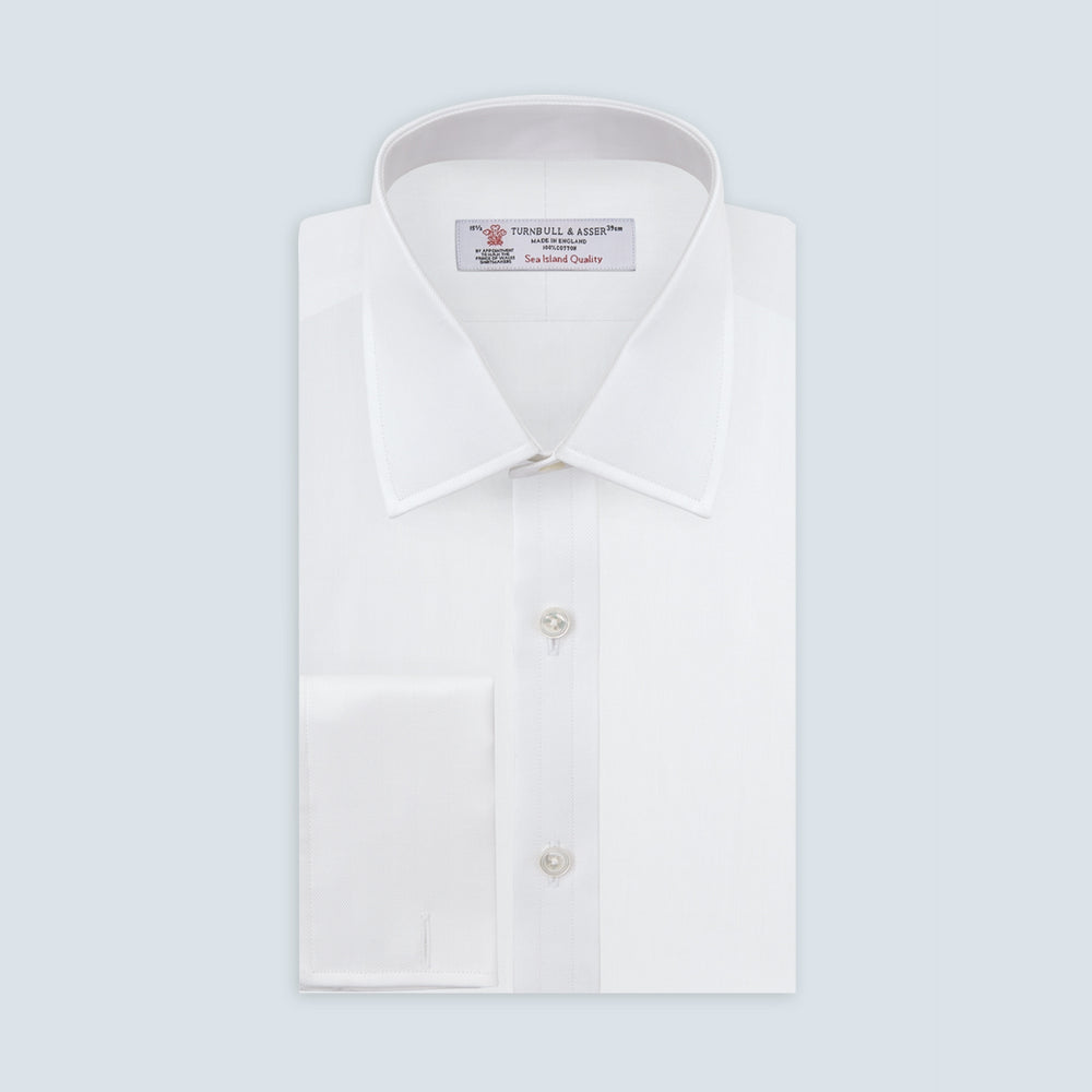 The Turnbull & Asser White Herringbone Sea Island Quality Cotton Shirt features a T&A collar, double cuffs, and chest pocket. Crafted for luxury with a classic design, it’s shown neatly folded against a light grey background—ideal for evening wear.