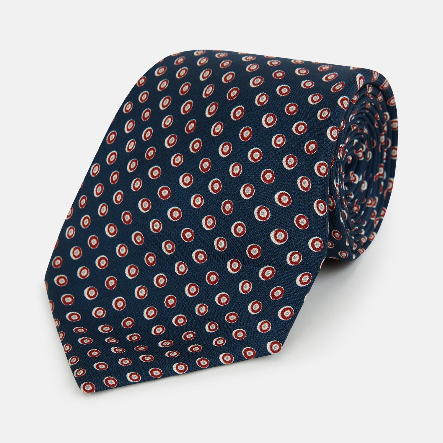 View 1 of product The Turnbull & Asser Navy and Red Circle Foulard Silk Tie, featuring a repeating red and white circular motif, is neatly rolled to display classic British craftsmanship.