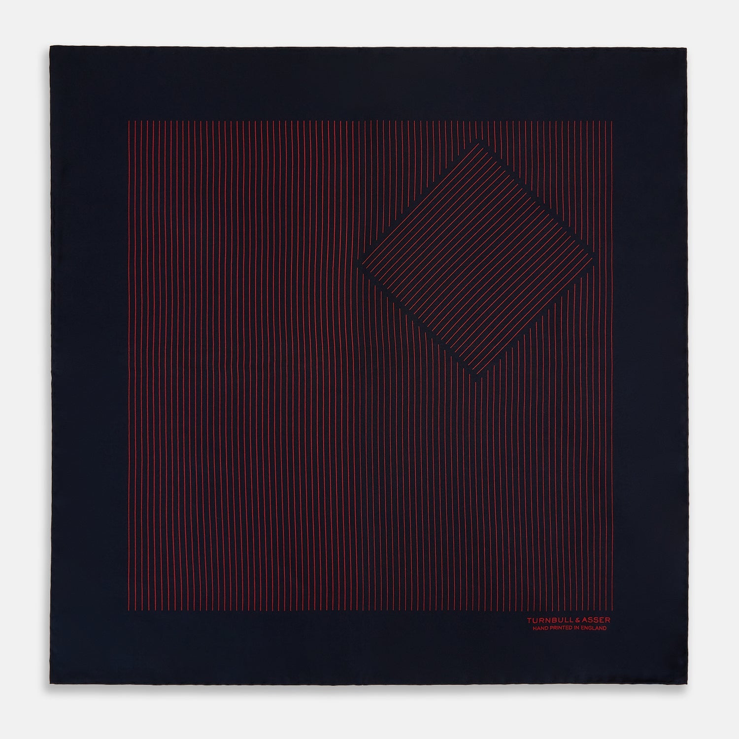 View 4 of product The Turnbull & Asser Navy and Red Stripe Silk Pocket Square features thin red stripes on navy silk, with "HAND PRINTED IN ENGLAND" in red at the corner. A folded corner shows its hand-rolled edges and refined design.