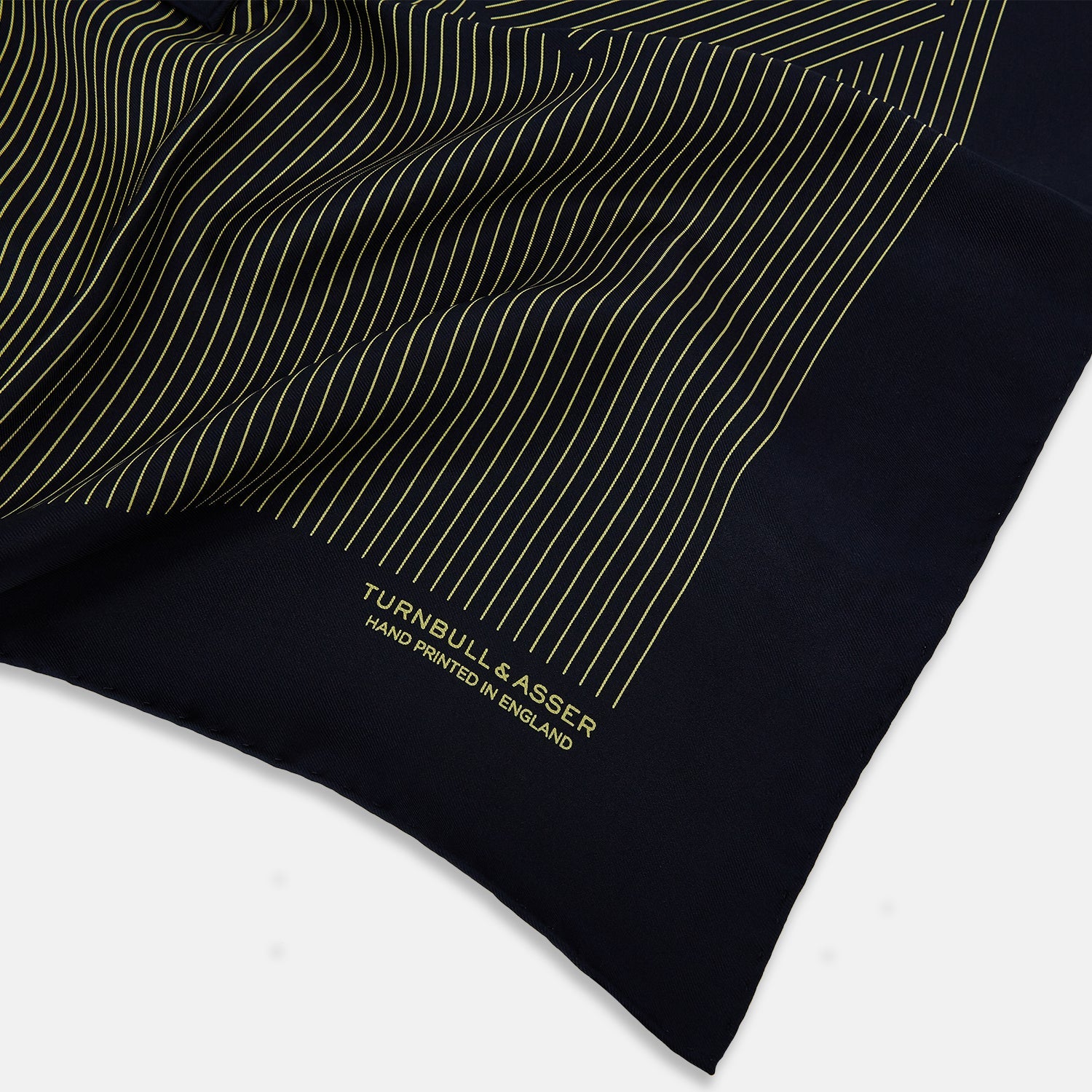 View 2 of product The Turnbull & Asser Navy and Gold Stripe Silk Pocket Square features light yellow vertical stripes, a folded corner, and "TURNBULL & ASSER HAND PRINTED IN ENGLAND" in gold at the bottom right.