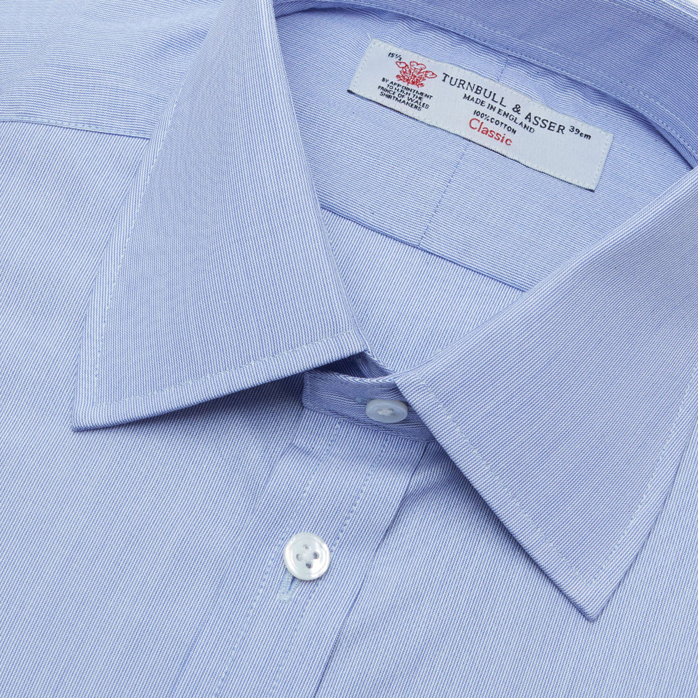 The Turnbull & Asser Blue Hairline Stripe Shirt features a T&A collar, 3-button cuffs, and white buttons. Made from light blue cotton with subtle stripes, it’s neatly folded with the label visible inside the collar on a plain white background.
