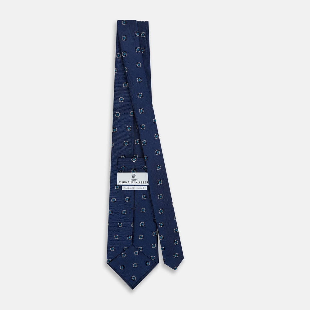 The Turnbull & Asser Navy Geometric Silk Tie showcases a repeating pattern of small light blue and white shapes on a navy base, neatly rolled and displayed against a white background.