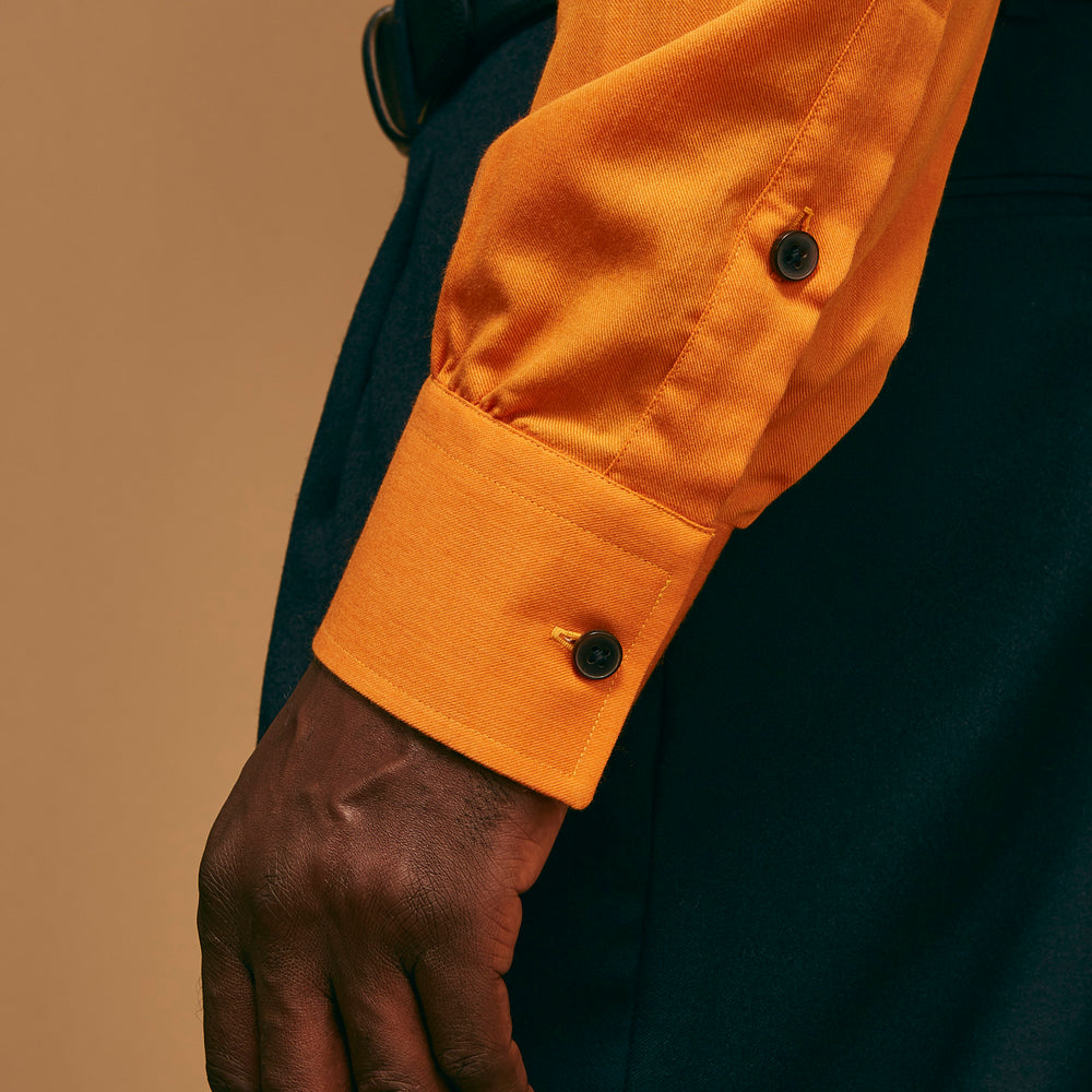 A man stands against a tan background, wearing a Turnbull & Asser Orange Weekend Fit Cotton & Wool Shirt with Derby Collar and 1 Button Cuffs, tucked into dark blue dress pants with a black belt and shoes. He has one hand in his pocket.